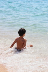 A boy is sitting in the ocean, looking out at the water in a beach.