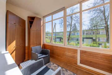 Interior of cozy balcony with wooden walls, sitting area with rug, armchair, wardrobe. Sunlight