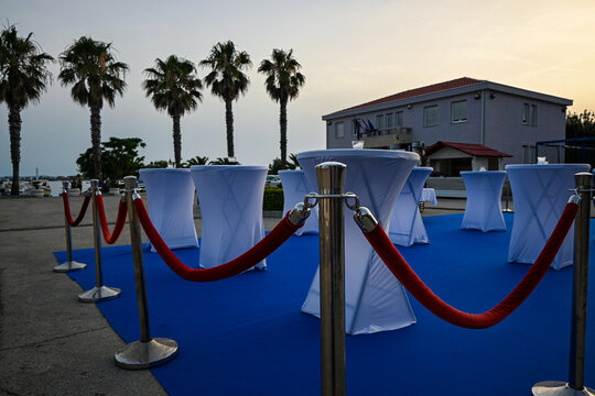 Vip area is set up outside for an event with white cocktail tables, red velvet ropes, and a blue carpet