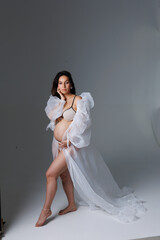 A beautiful pregnant brunette woman in a white flying transparent negligee, dressing gown. A pretty girl with a bare stomach in lingerie stay barefoot on a grey background. Smile, portrait