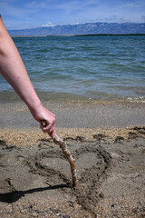Person is drawing a heart shape in the sand on a beach with a stick