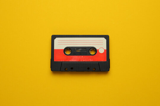 A cassette tape is laying on a yellow background. The tape is old and worn, with a red and white stripe. The tape is placed on a yellow surface, which gives the image a nostalgic and vintage feel