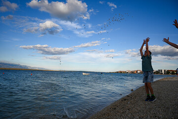 Young boy throwing stones into a calm blue sea on a summer day with white clouds in the sky