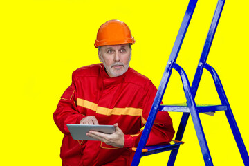 Senior male worker holding tablet pc leans on a blue ladder. Isolated on yellow.