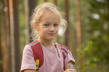 A young girl wearing a pink shirt and red backpack drinks from a colorful water bottle decorated with watermelon slices, standing in a sunlit forest.