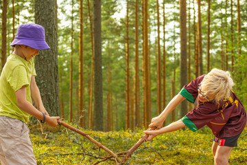 Two boys, one in a green shirt and purple hat and the other in a maroon shirt, energetically play with a large stick in a forest, enjoying the outdoors and each others company.