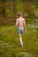 A young girl with a braided ponytail, wearing a pink shirt and floral denim shorts, walks through a lush forest, surrounded by greenery.