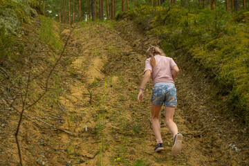 A young girl with a braided ponytail, wearing a pink shirt and floral denim shorts, walks through a lush forest, surrounded by greenery.