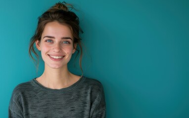 Fototapeta premium A young woman with brown hair tied up in a bun smiles at the camera while standing in front of a teal wall