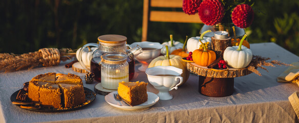 Tea setting with handmade pie. Beautiful view of garden wooden furniture with basket, soft blanket and burning candles. Outdoors fall family party, cozy autumn interior. Pumpkins, red dahlia banner