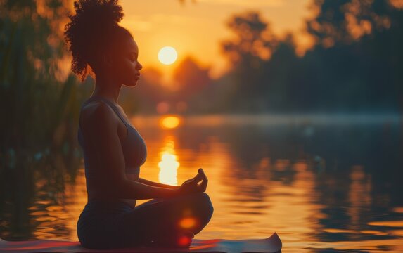 A young black woman with a ponytail sits in a yoga pose on a mat at the edge of a lake. The sun sets behind her, casting a warm glow over the scene