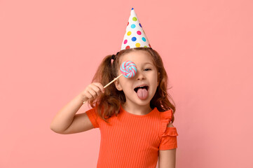 Cute little girl in holiday cap covering one eye with lollipop and showing tongue on pink background