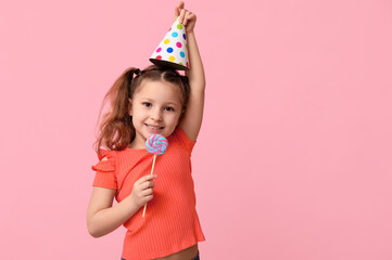 Cute little girl in party cap with lollipop on pink background