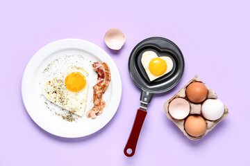 Composition with heart-shaped fried egg in frying pan and plate with fried egg and bacon on lilac background
