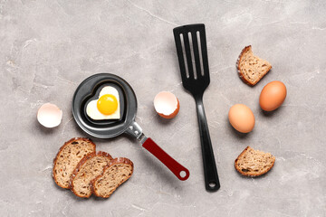 Composition with heart-shaped fried egg in frying pan, eggs, eggshell, spatula and bread on grunge background