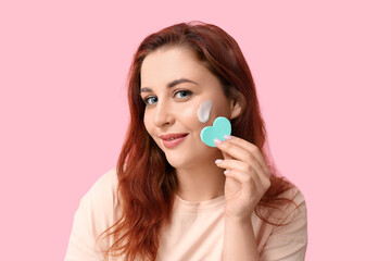 Beautiful young woman with heart-shaped makeup sponge on pink background