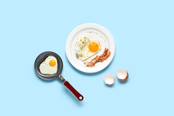 Composition with eggshell, heart-shaped fried egg in pan and in plate on blue background
