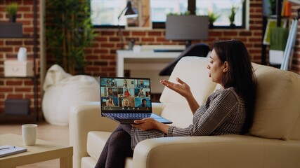 Teleworking team leader sitting on couch in cozy apartment, chatting with coworkers during teleconference meeting. Indian business manager at home holding online videocall with workers, camera B