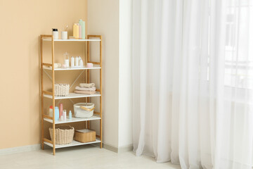 Rack with bottles of cosmetic products, towels and wicker baskets near beige wall