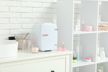 Small cosmetic refrigerator, bottles and jars of cosmetic products on white chest of drawers