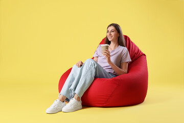 Smiling woman with paper cup of drink sitting on red bean bag chair against yellow background