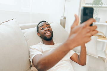 Happy African American Man Sitting on Black Sofa, Using Mobile Phone and Typing Message in Modern...