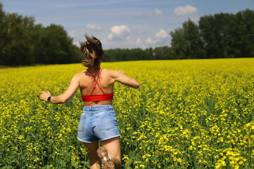 Adult happy  woman in a red top and denim shorts runs through a yellow rapeseed field. Motion blur effect.