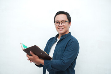 Young Asian man holding books smiling happy and positive while reading a book. Asian student flipping a book page over white background