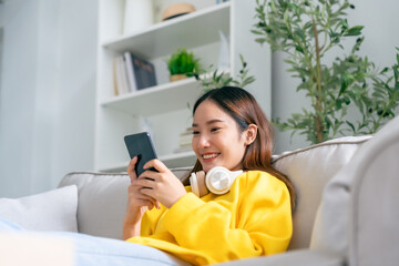 Happy young asian woman  wearing headset and relaxing on comfortable couch at home, texting...