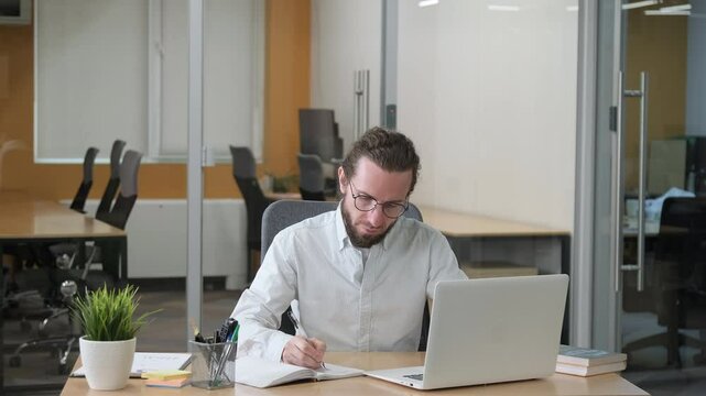 A professional employee is seen deeply engaged with his tasks, writing notes beside his laptop. Office worker busy on his desk. Planning.