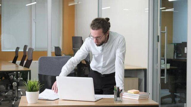 Business man standing in his office while working on his laptop. A businessman conducts detailed research, comparing digital and printed financial data.