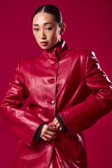 Fashionable asian brunette in red leather jacket posing against red background with hands on hips