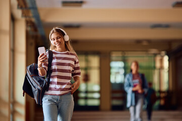 Happy high school student using cell phone while listening music on headphones in hallway.