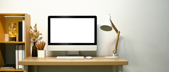 Front view of blank screen computer desktop, lamp and books on wooden table against white wall