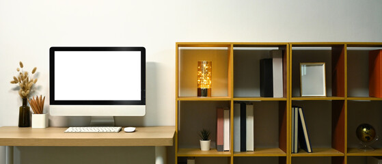 Blank screen computer desktop, lamp and books on wooden table near bookshelf