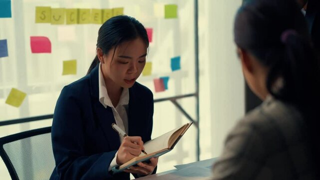 Businesswoman discusses tasks and takes notes during a meeting with a colleague in a modern office. Sticky notes with the word success are visible in the background