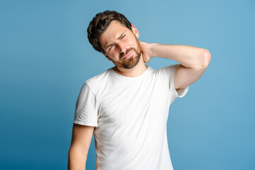 Portrait of young man feeling pain holding his neck with his hand, over blue background