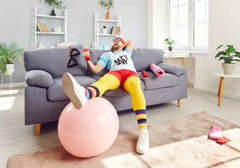Portrait of funny young man wearing sportswear sitting on sofa with sport equipment and dumbbells...