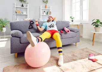 Gordijnen Portrait of funny young man wearing sportswear sitting on sofa with sport equipment and dumbbells in hands in the living room at home. Fitness, workout sport and home training concept. © Studio Romantic