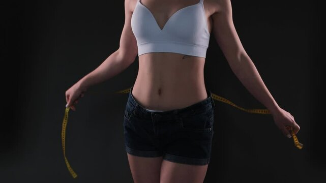 A slender girl in a beige white top and short black denim shorts measures her waist and hips with a measuring tape on a dark background. The concept of weight loss and healthy lifestyle.
