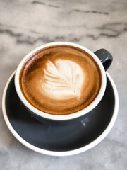Coffee Latte art with white coffee art flower in brown foam in a black coffee cup on a black saucer on a marble table