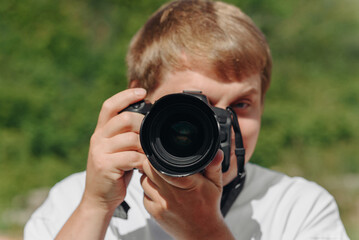 dslr camera in hands, young adult caucasian photographer in white t-shirt at work outdoors in sunny summer day, looking at camera and taking picture