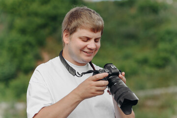 dslr camera in hands, young adult caucasian photographer in white t-shirt at work outdoors in sunny summer day, watching photos at display and smiling