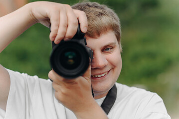 dslr camera in hands, young adult caucasian photographer in white t-shirt at work outdoors in sunny summer day, looking at camera and taking picture and smiling