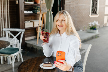 young beautiful blond caucasian woman sitting at table in outdoor terrace of cafe in summer day with chocolate cheesecake and strawberry cocktail, businesswoman holding smartphone and smiling