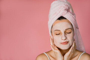 A young woman in a pink spa standing in towel and mask looking relaxed and putting mask on 