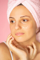 A woman with smooth skin standing in the bathroom doing her spa procedures, using eyepatches and a pink towel 