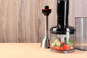 Hand blender kit with fresh vegetables and parsley on beige table against wooden background, space...