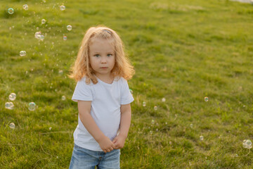 Fototapeta premium Cute blonde kid with curly hair walks on the lawn with soap bubbles in summer