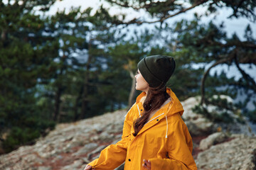 A woman in a vibrant yellow raincoat standing majestically on top of a mountain surrounded by lush green trees